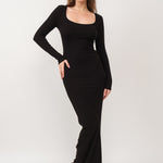 A model wearing a black ribbed maxi dress against a white wall.