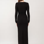 A model wearing a black ribbed maxi dress against a white wall.