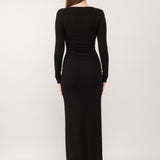 A model wearing a black ribbed maxi dress against a white wall. 