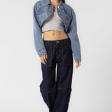 A model wearing a cropped denim jacket full body image against a white wall. 