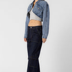 A model wearing a cropped denim jacket sideview full body image against a white wall.