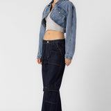 A model wearing a cropped denim jacket sideview full body image against a white wall. 