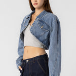 A model wearing a cropped denim jacket with a sideview against a white wall.