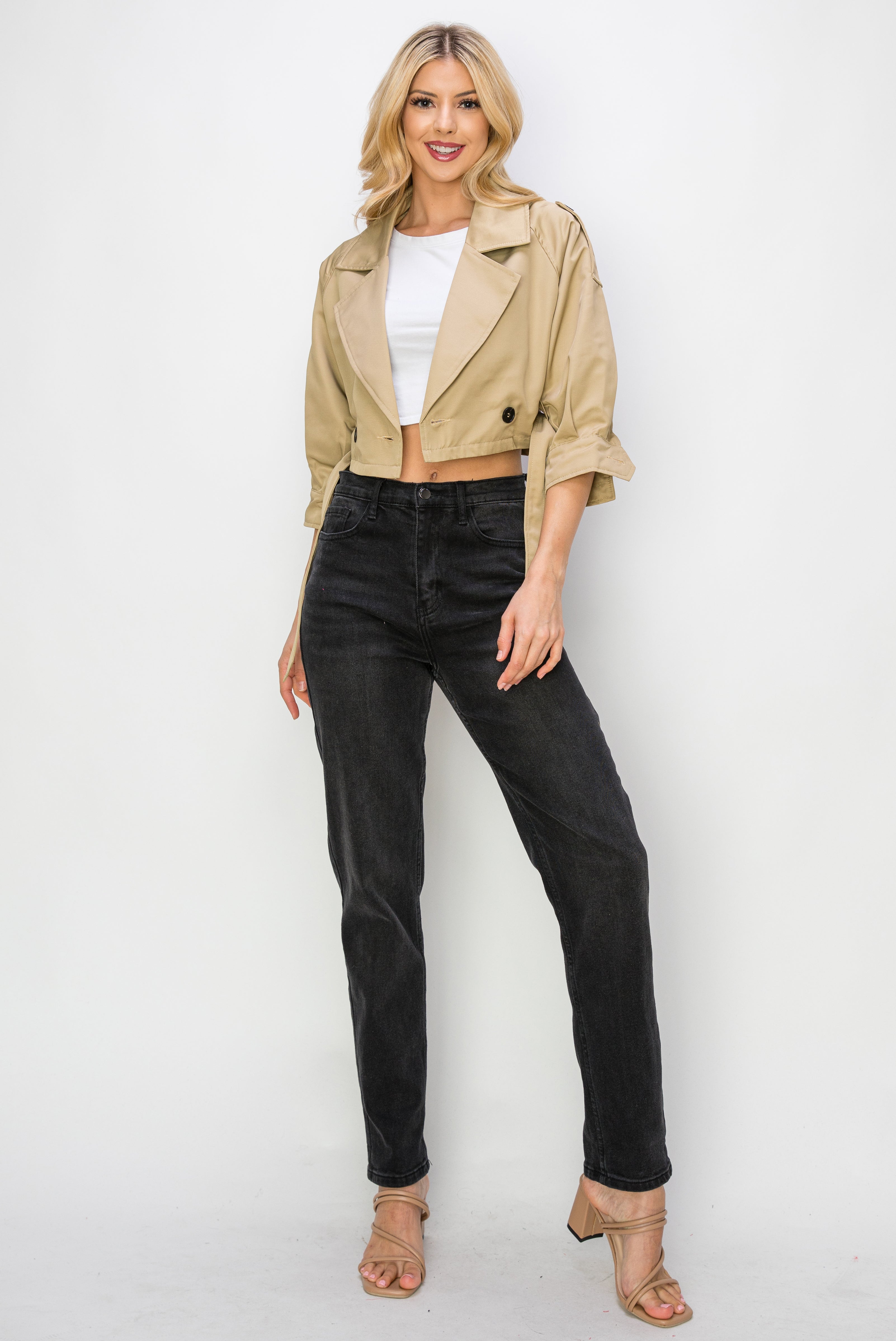 A model wearing a cropped trench jacket opened at the front against a white wall.