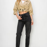 A model wearing a cropped trench jacket full body image against a white wall. 