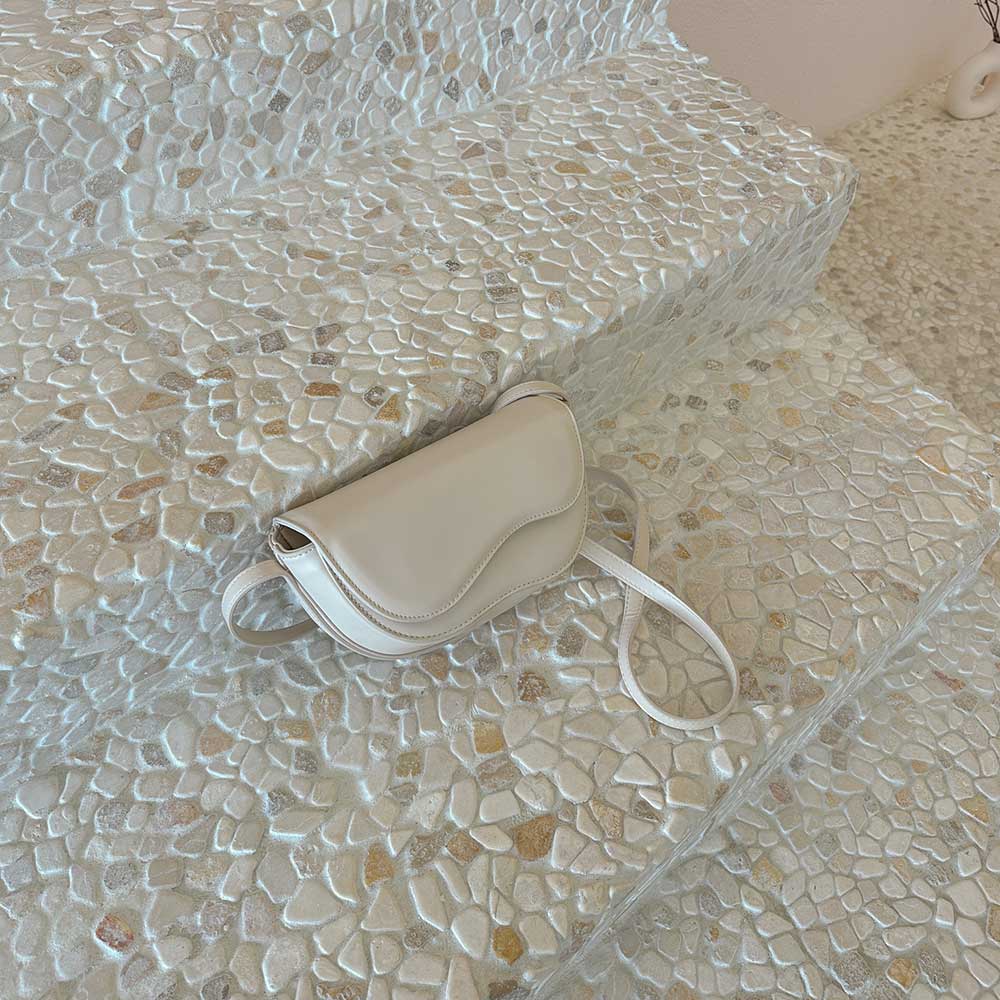 A still image of a small nude vegan leather crossbody bag with a wavy front flap closure laying against a stone background.