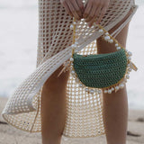 A model wearing a small crochet straw top handle bag with seashell details along the handle while standing on the beach.