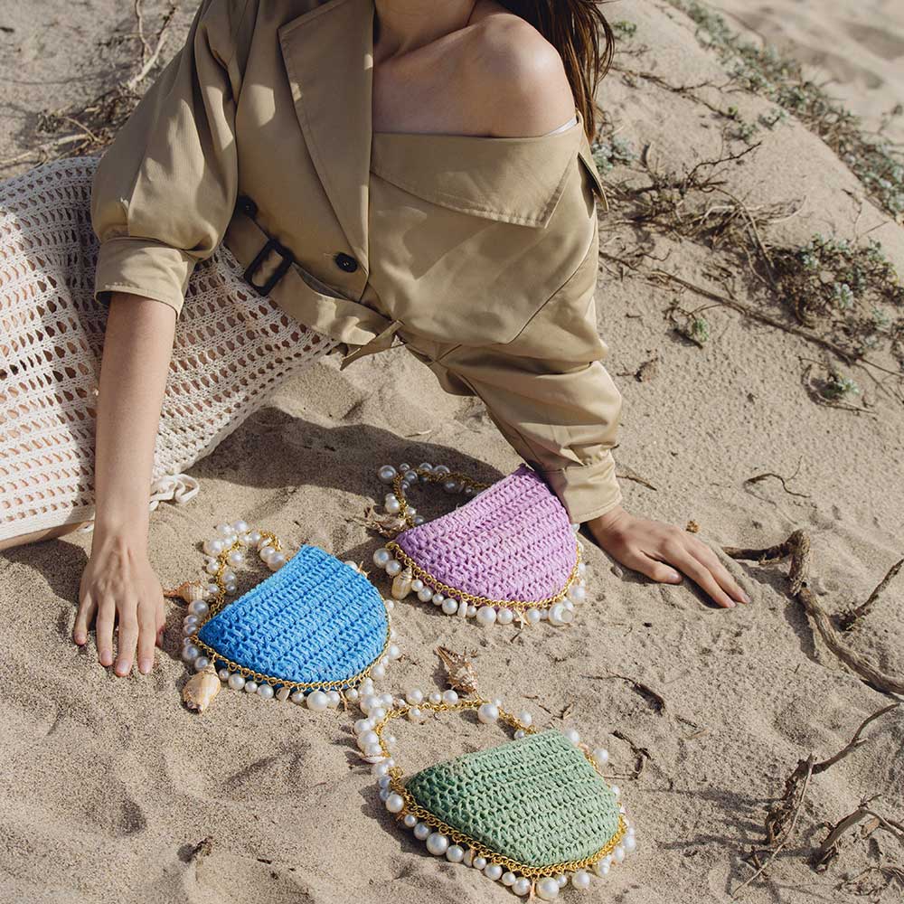A model with three crochet straw top handle bag with seashell details along the handle while laying in the sand.