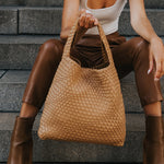 A model holding a large woven recycled vegan leather shoulder bag sitting on stairs. 