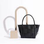 A still image of a small black woven wide strap tote bag against a white wall with wood props. 