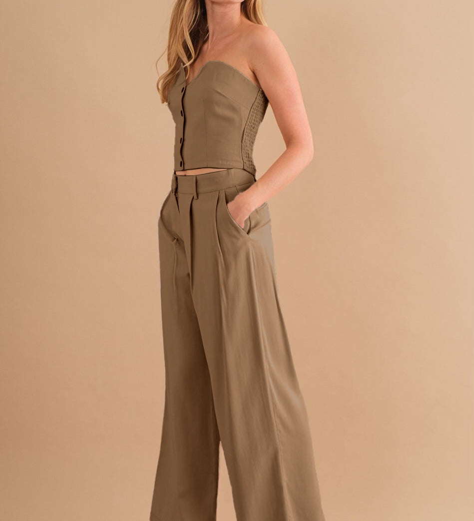 A model wearing a tan two piece strapless top and pant set against a tan wall. 
