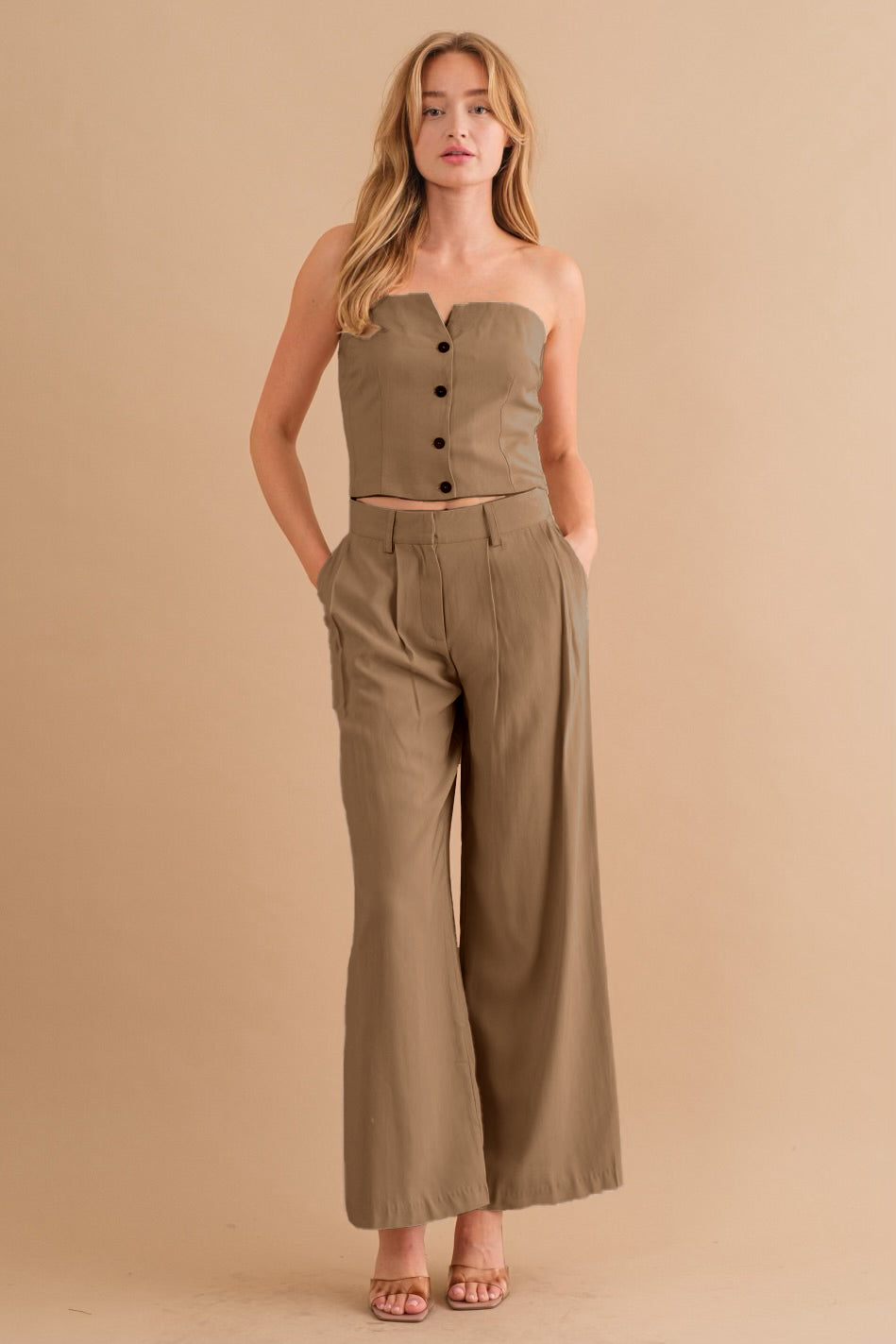 A model wearing a two piece strapless top and pant set against a tan wall. 