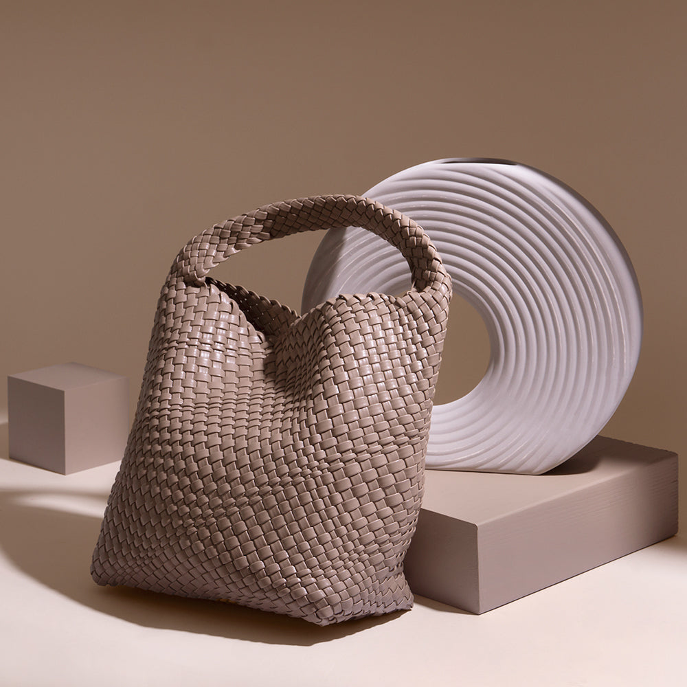 A still image of a large woven vegan leather shoulder bag against a brown wall with a white vase.