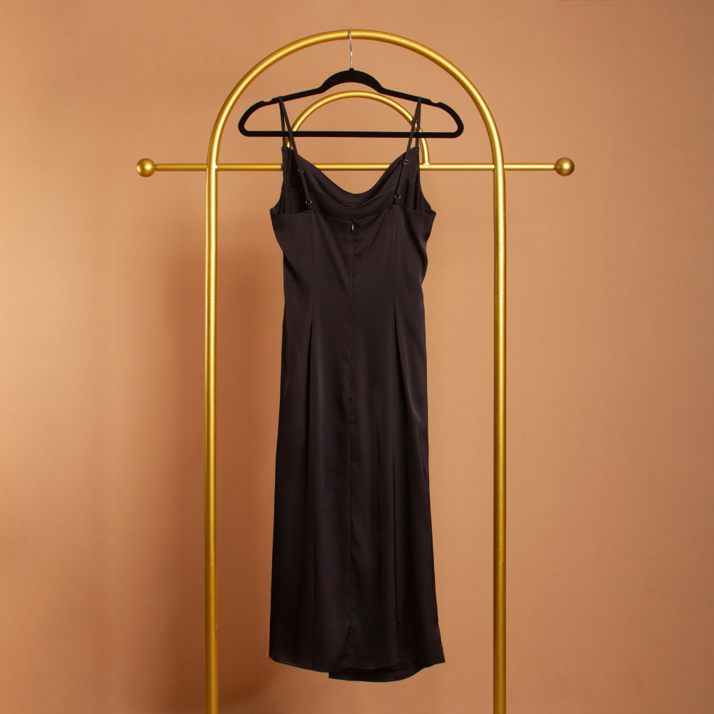 A black satin cowl neck slip dress backside view on a hanger with an orange background. 