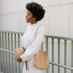 A model wearing a pleated vegan leather shoulder bag while holding a zip pouch against a railing outside. 