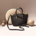 A still image of a black woven vegan leather crossbody bag with a wrapped handle against a stone prop. 