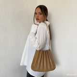 A model wearing a taupe pleated vegan leather shoulder bag against a white wall. 