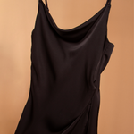 A detail image of a black satin cowl neck slip dress against an orange background. 