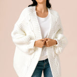 A model wearing a white knitted cardigan against a tan background. 