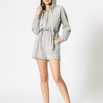 a model wearing a grey terry hooded romper against a white wall.