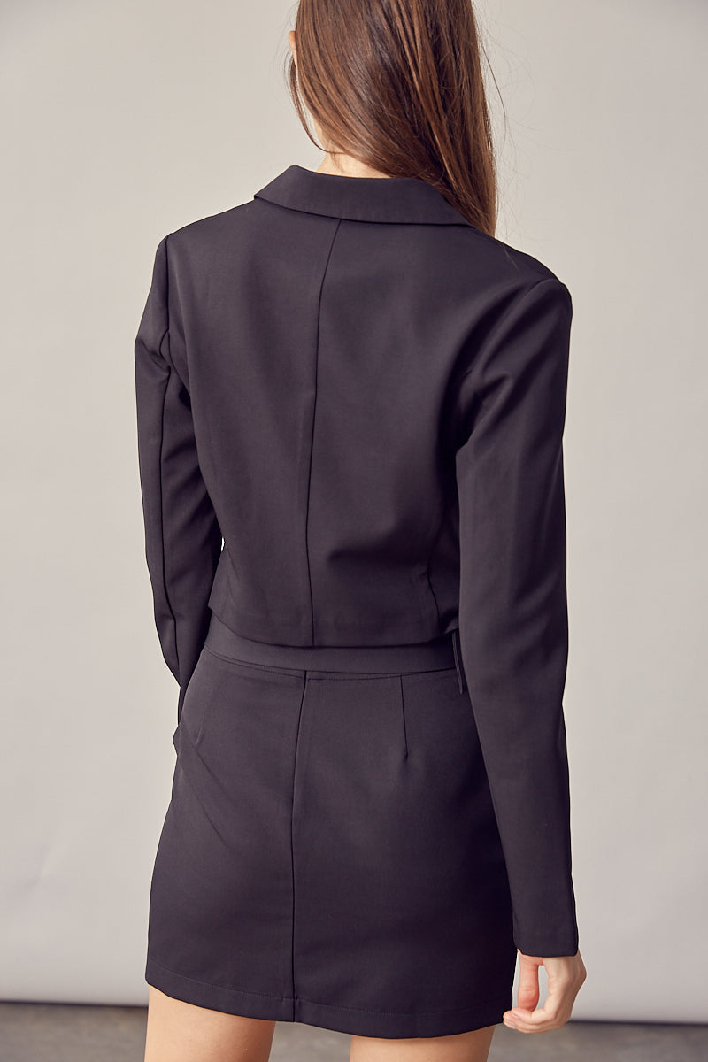 A model wearing a black double breasted cropped jacket backside view against a white wall. 