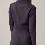 A model wearing a black double breasted cropped jacket backside view against a white wall. 