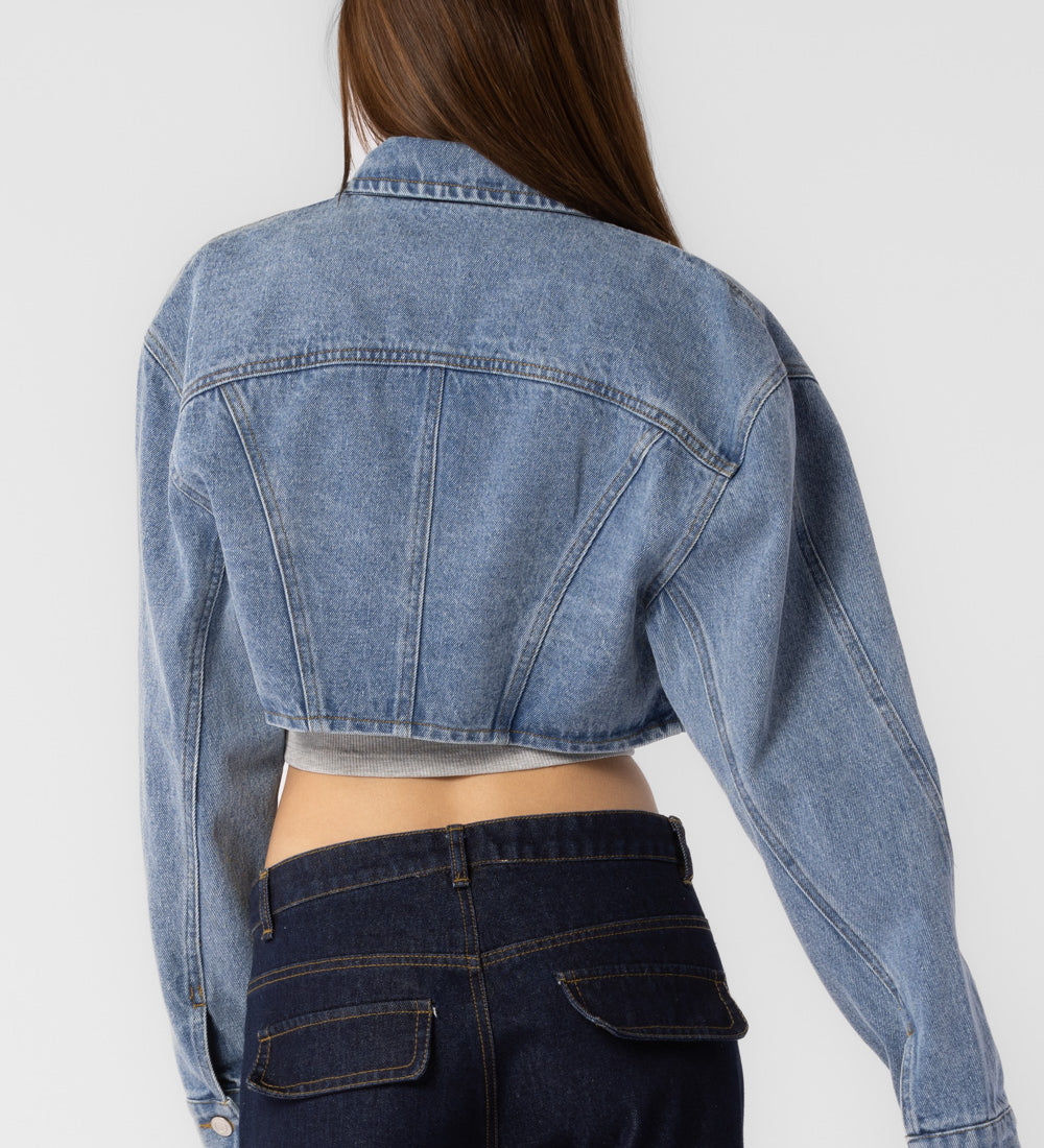 A model wearing a cropped denim jacket with backside view against a white wall. 