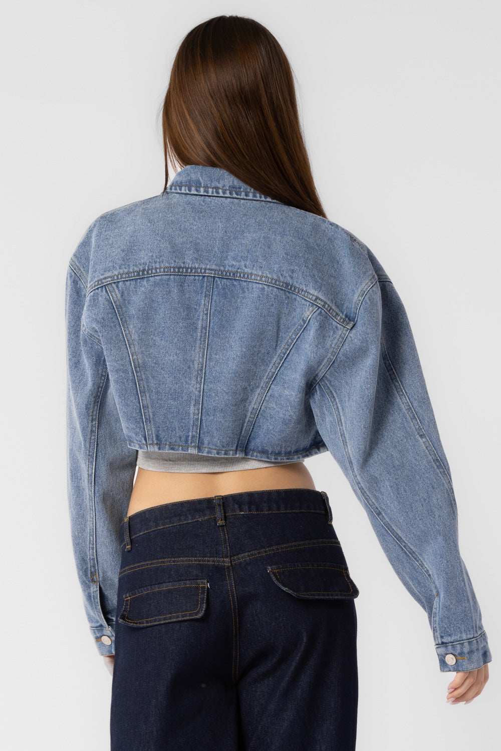 A model wearing a cropped denim jacket with backside view against a white wall. 