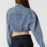 A model wearing a cropped denim jacket with backside view against a white wall. 