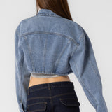 A model wearing a cropped denim jacket with backside view against a white wall. 