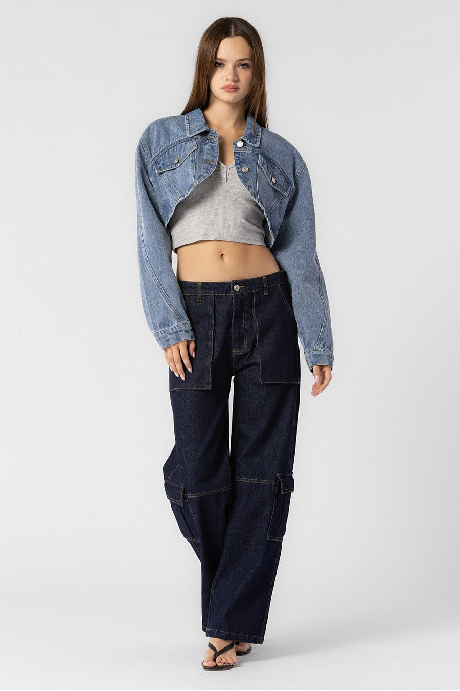 A model wearing a cropped denim jacket full body image against a white wall. 