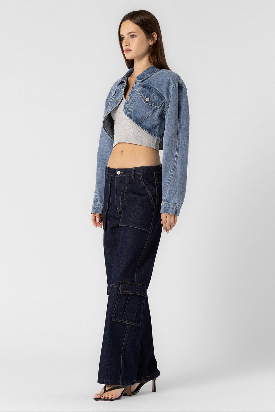 A model wearing a cropped denim jacket sideview full body image against a white wall. 