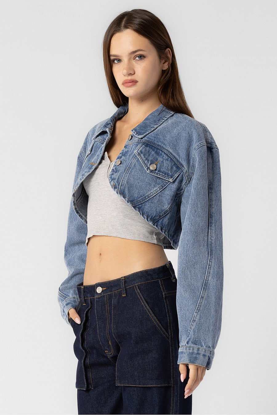 A model wearing a cropped denim jacket with a sideview against a white wall.