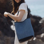 A model wearing a large denim tote bag with a double knotted handle while on the beach.