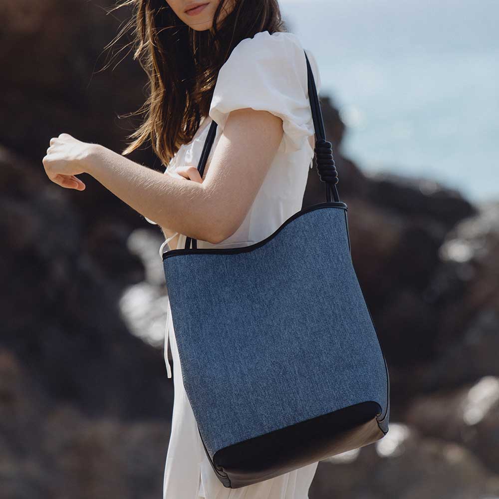 A model wearing a large denim tote bag with black trimming while on the beach.