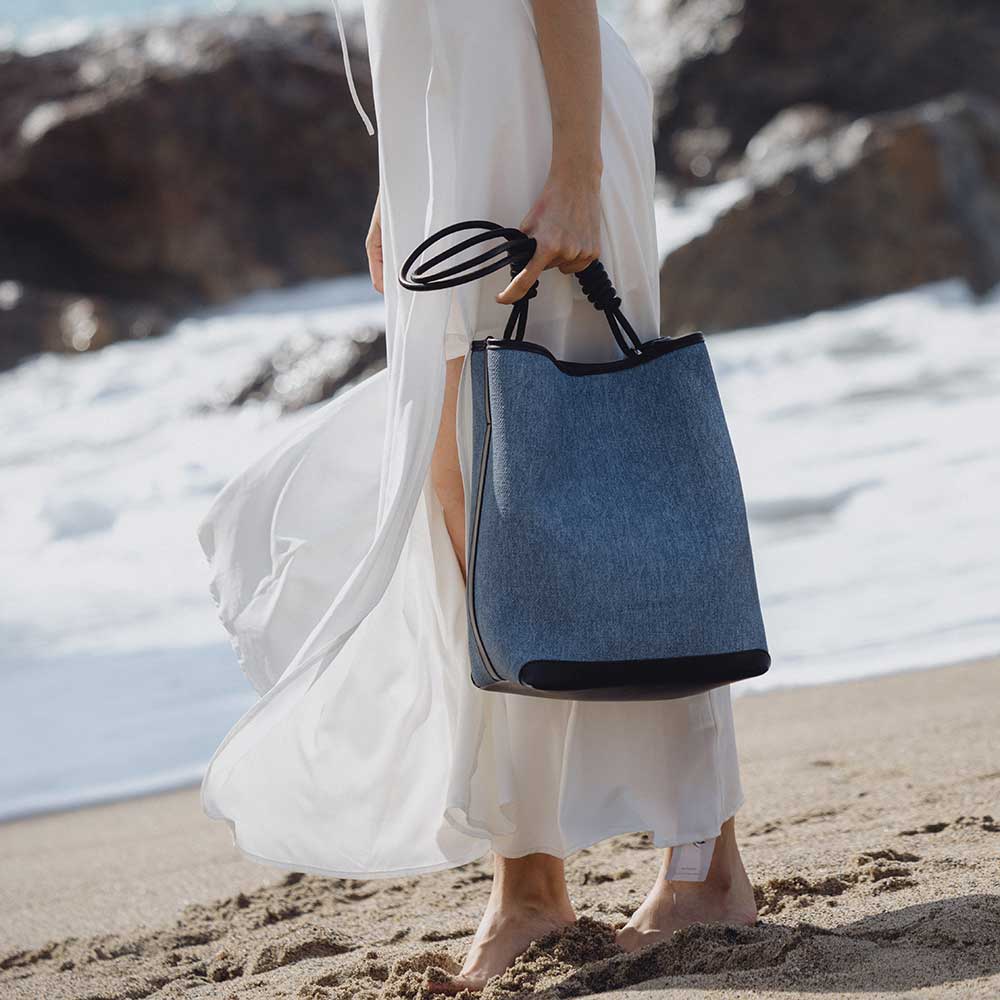 A model wearing a large denim tote bag with black trimming while on the beach.