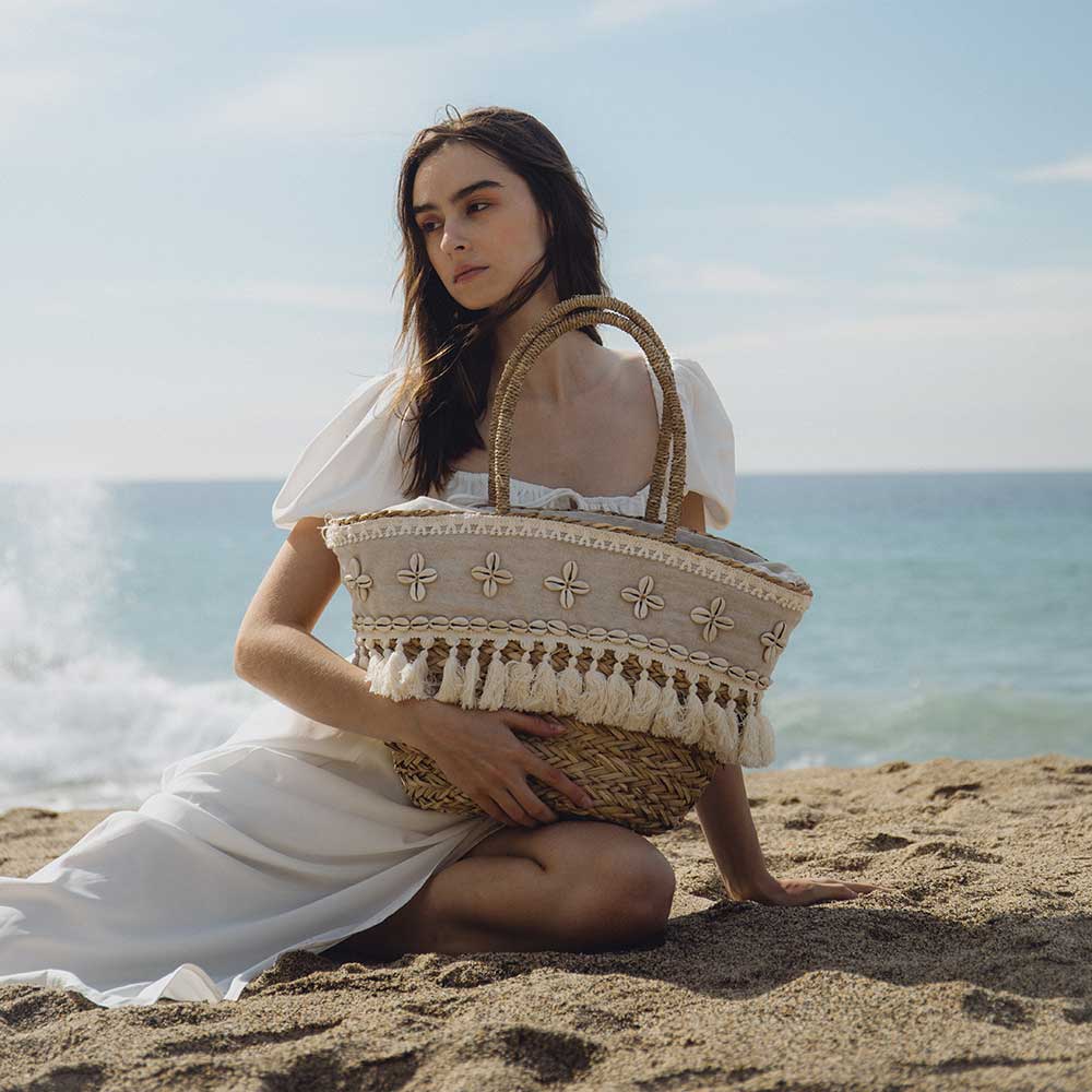 A model with a large straw woven tote bag with seashell details with a drawstring closure sitting on the beach.