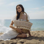 A model with a large straw woven tote bag with seashell details with a drawstring closure sitting on the beach.