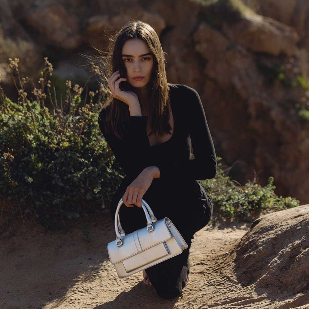 A model wearing a small silver metallic vegan leather rectangle shaped top handle bag while sitting outside.