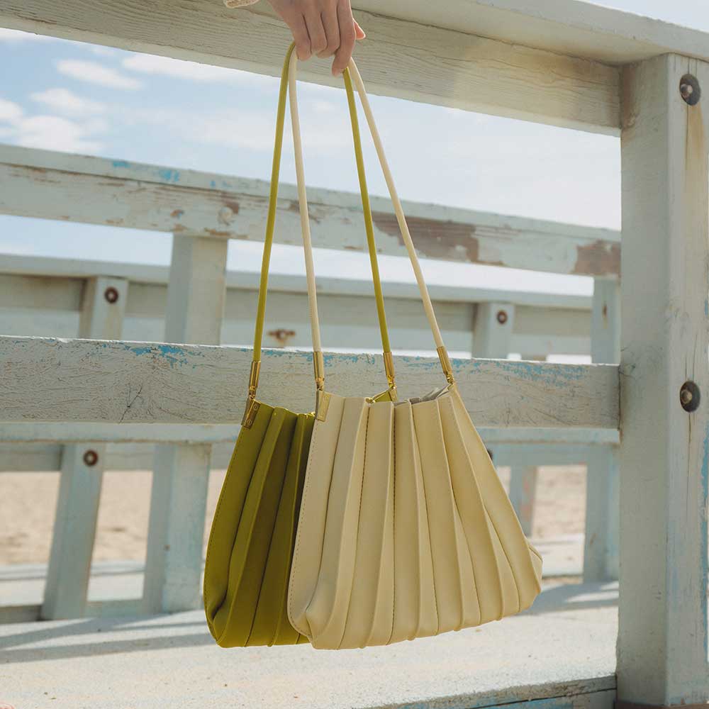 A still image of two medium pleated vegan leather shoulder bags held by a model on the beach.