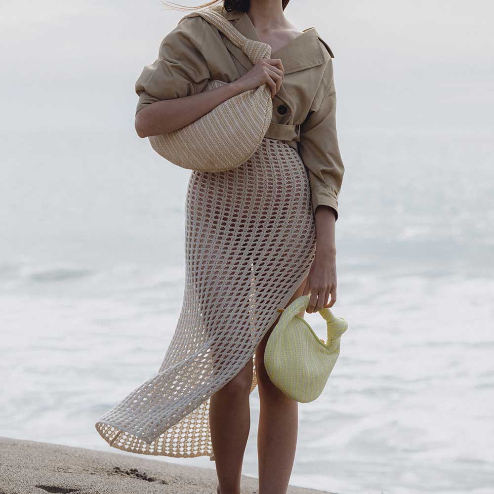 A model wearing two straw woven handbags with knot handles while standing on the beach 