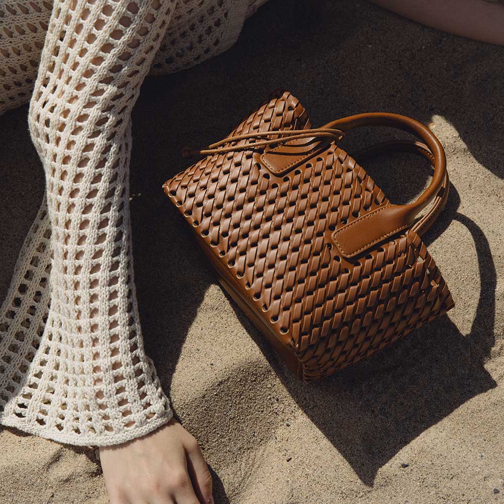 A still image of a small saddle woven vegan leather top handle bag with a drawstring closure laying in sand.
