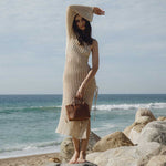 A model wearing a small saddle woven vegan leather top handle bag with a drawstring closure while standing on the beach.