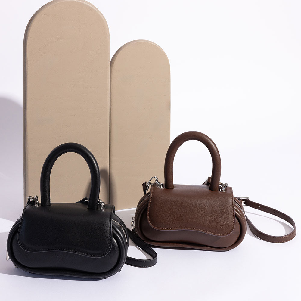 A still image of two oval shaped crossbody handbags with tan props in the background. 