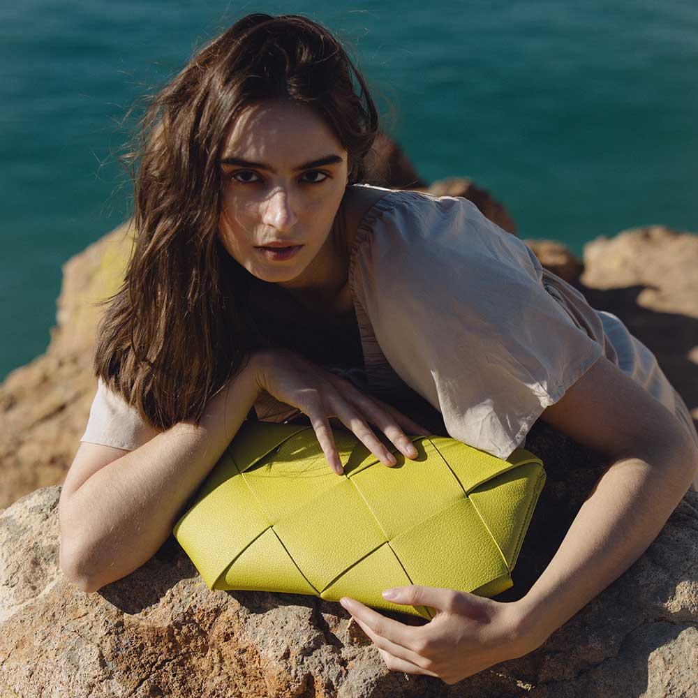 A model wearing a small lime woven vegan leather clutch with a crossbody strap while laying on rocks.