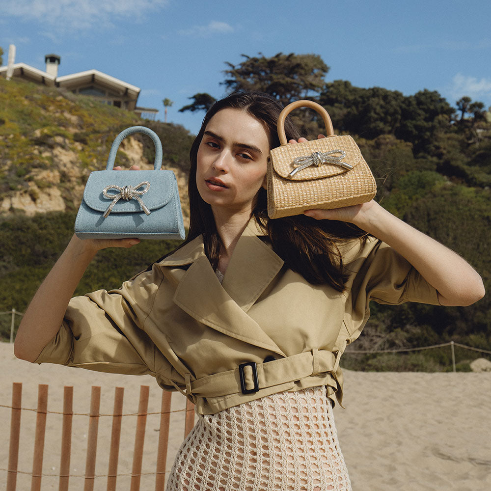 Model holding two small bags with rhinestone bows in a beach setting.