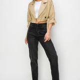 A model wearing a cropped trench jacket opened at the front against a white wall.