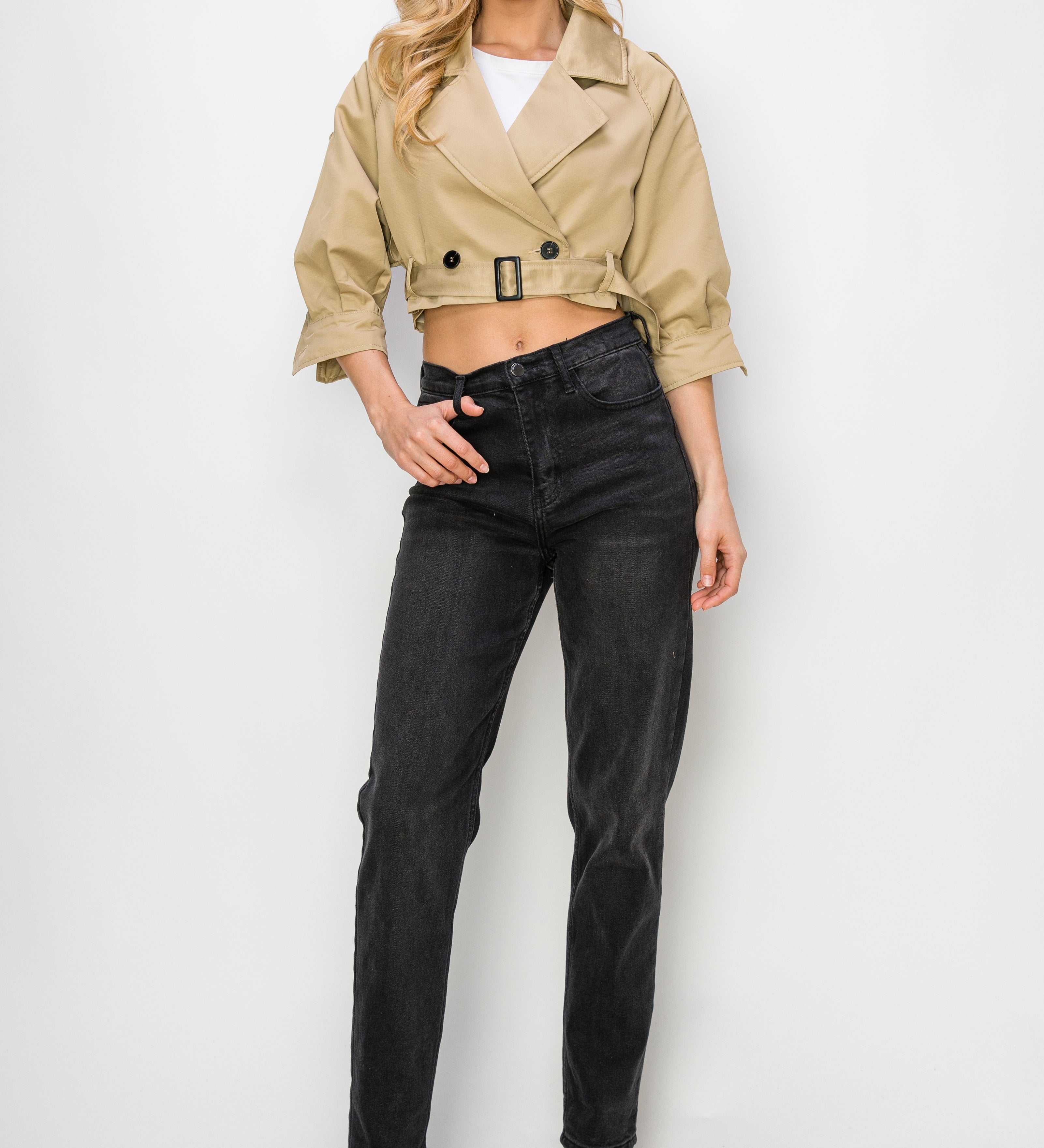 A model wearing a cropped trench jacket full body image against a white wall. 