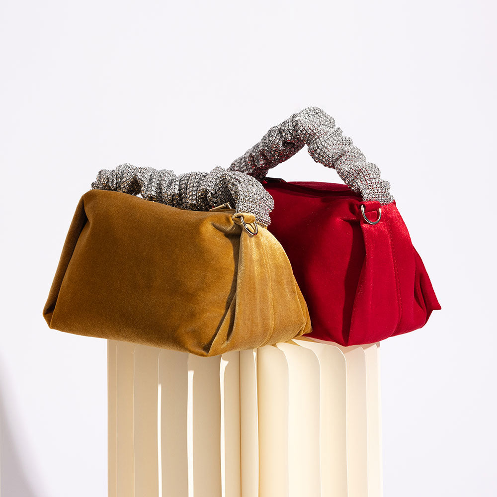 A still image of two medium velvet top handle bags against a white background on a column. 