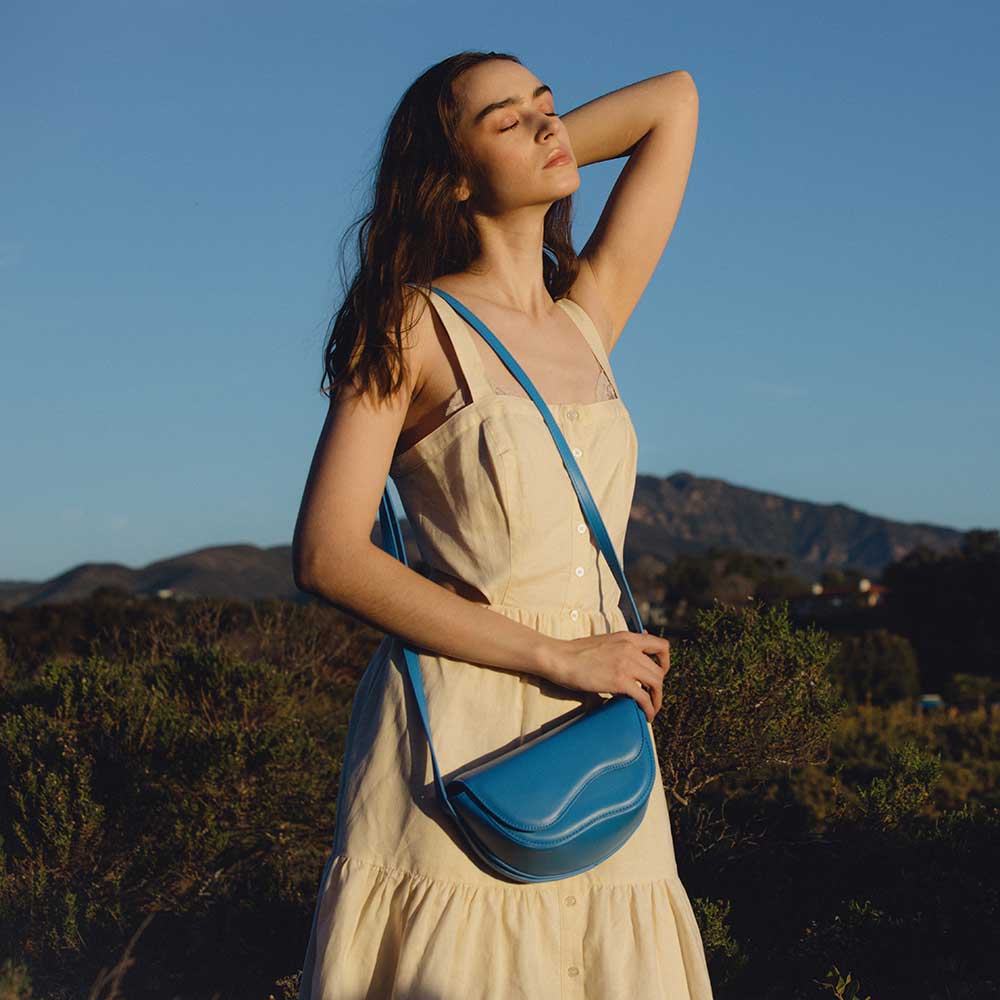 A model wearing a small blue vegan leather crossbody bag with a wavy front flap closure.